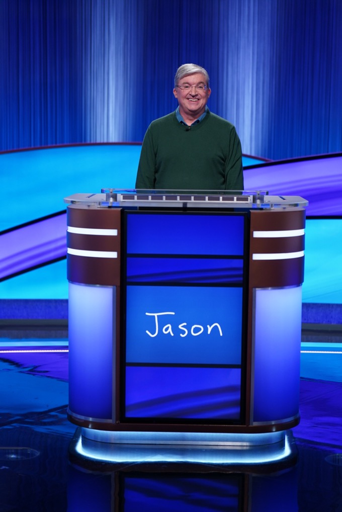 A man in a green sweater stands behind a blue podium with 'Jason' on it, smiling on a game show set with blue lighting and curtains.