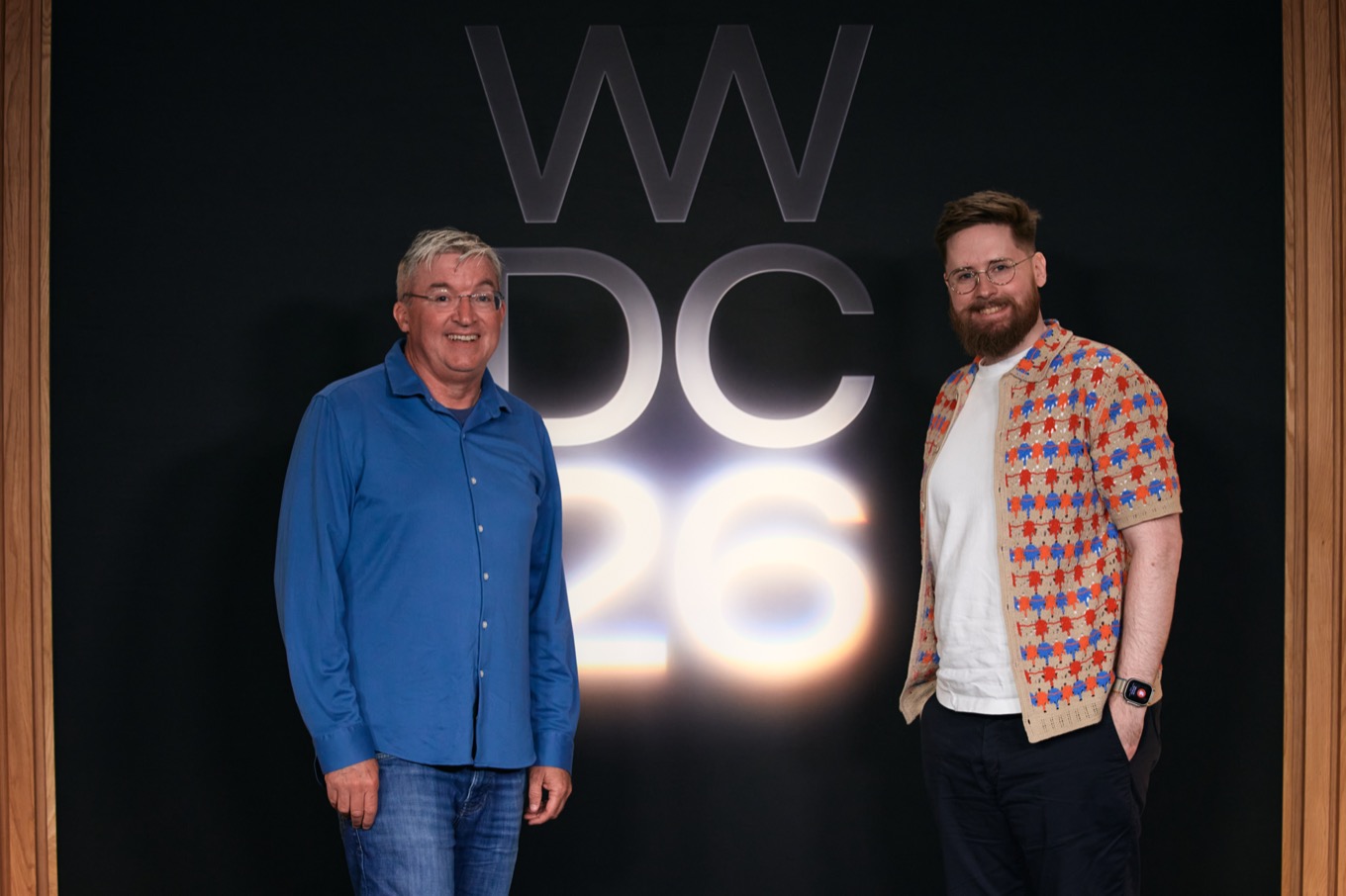Two men posing in front of a black backdrop with 'WWDC 26' in illuminated letters. One wears a blue shirt and jeans, the other a patterned shirt over a white tee and dark pants.